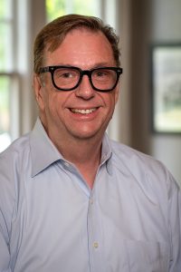 Photograph of a smiling middle-aged man wearing black glasses and a light blue dress shirt, standing indoors with natural light.