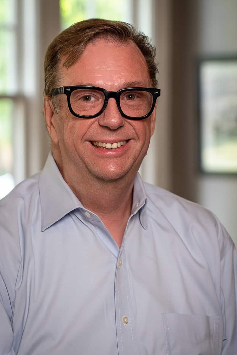 Photograph of a smiling middle-aged man wearing black glasses and a light blue dress shirt, standing indoors with natural light.